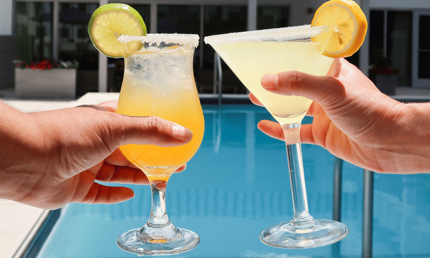 Two hands holding cocktails with lime and lemon by a pool.