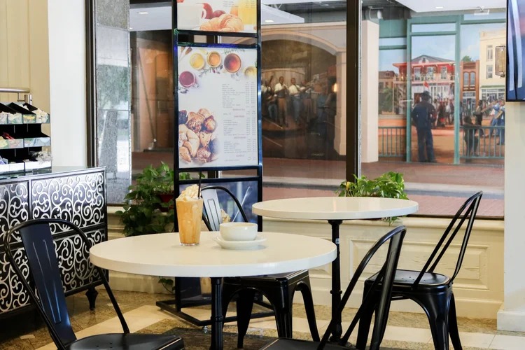 Cozy cafe interior with round white tables and black metal chairs; menu board with drinks and pastries; iced coffee and cup on table; decorative mural in background.
