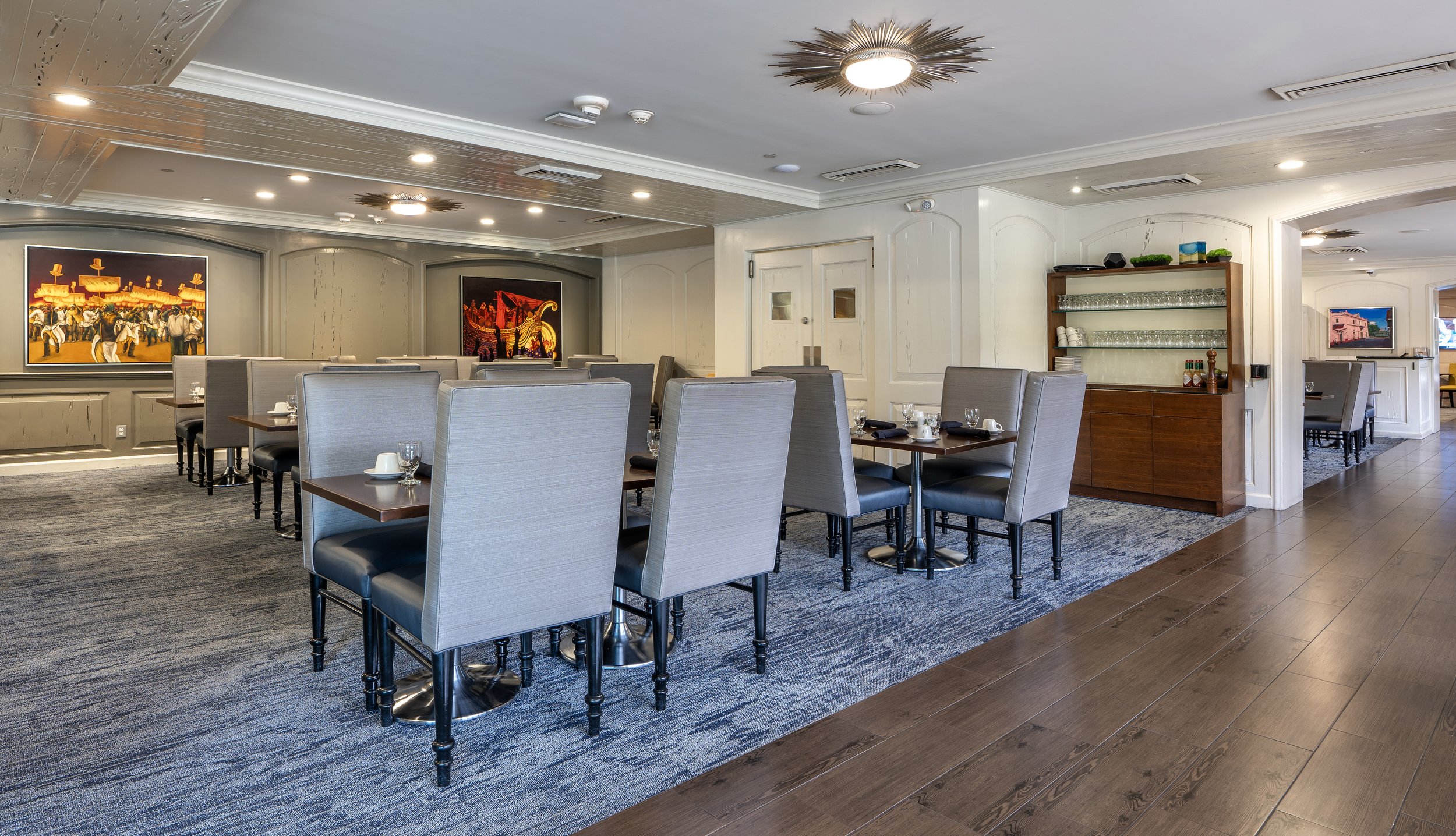Interior of a modern restaurant with gray upholstered chairs, blue carpet, wooden flooring, and decorative wall paintings.