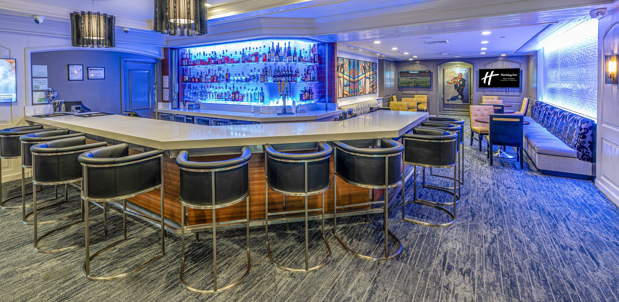 Modern hotel bar with a central counter, black bar stools, and a well-stocked liquor display with blue backlighting. Lounge seating area with couches and chairs, wall art, and a flat-screen TV.