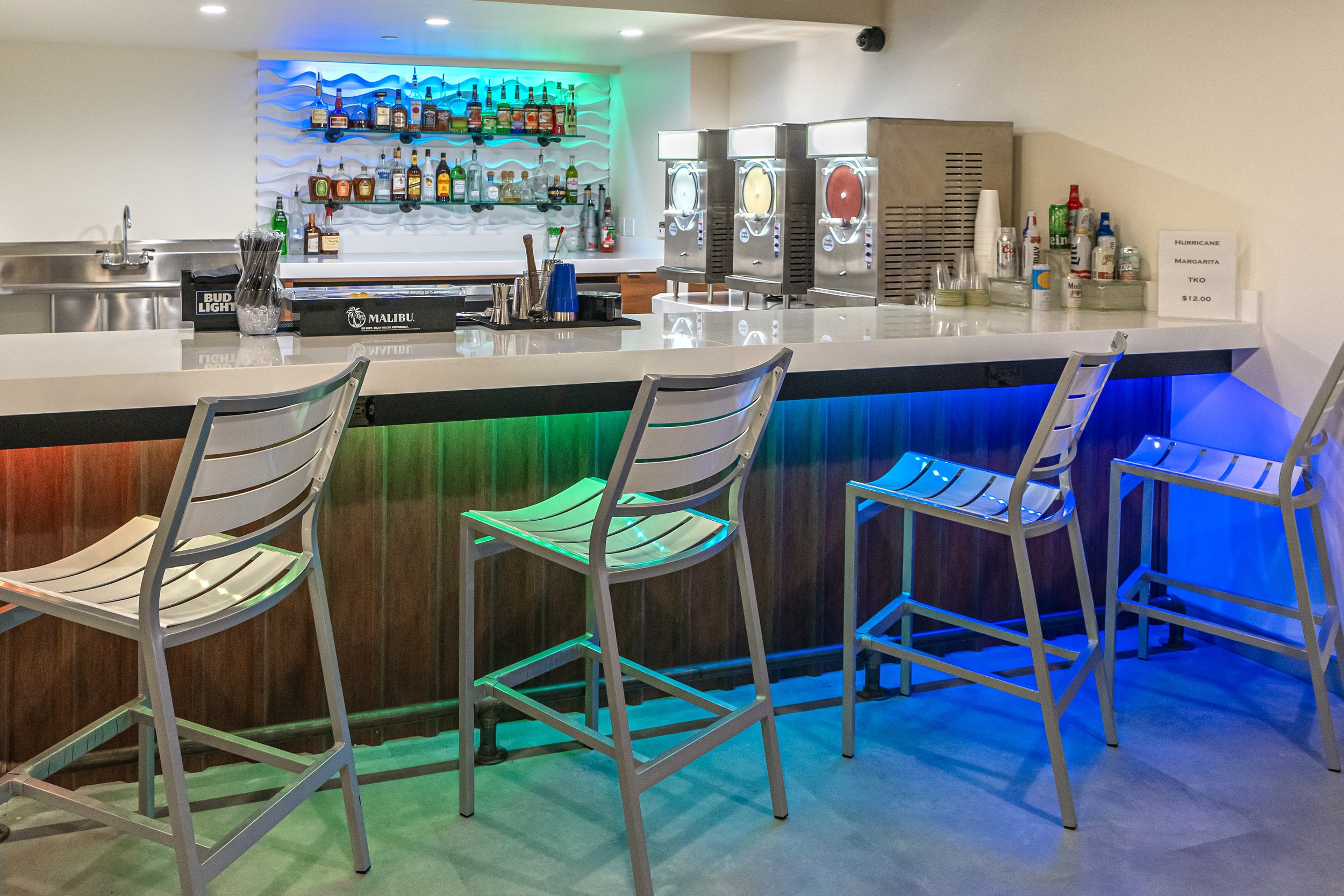 Modern bar with colorful LED lights, metal bar stools, bottles on shelves, and frozen drink machines.