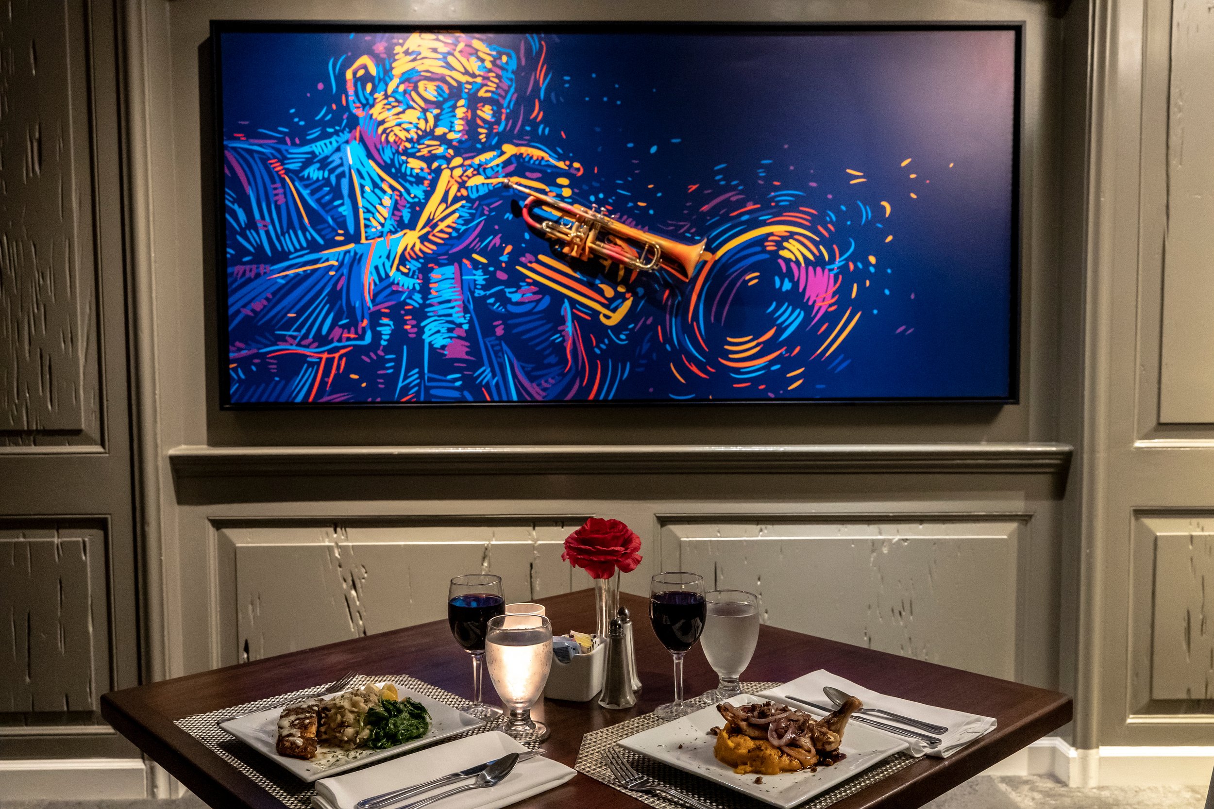 A dining table set for two with plates of gourmet food and wine glasses, beneath an artistic painting of a jazz musician playing a trumpet.