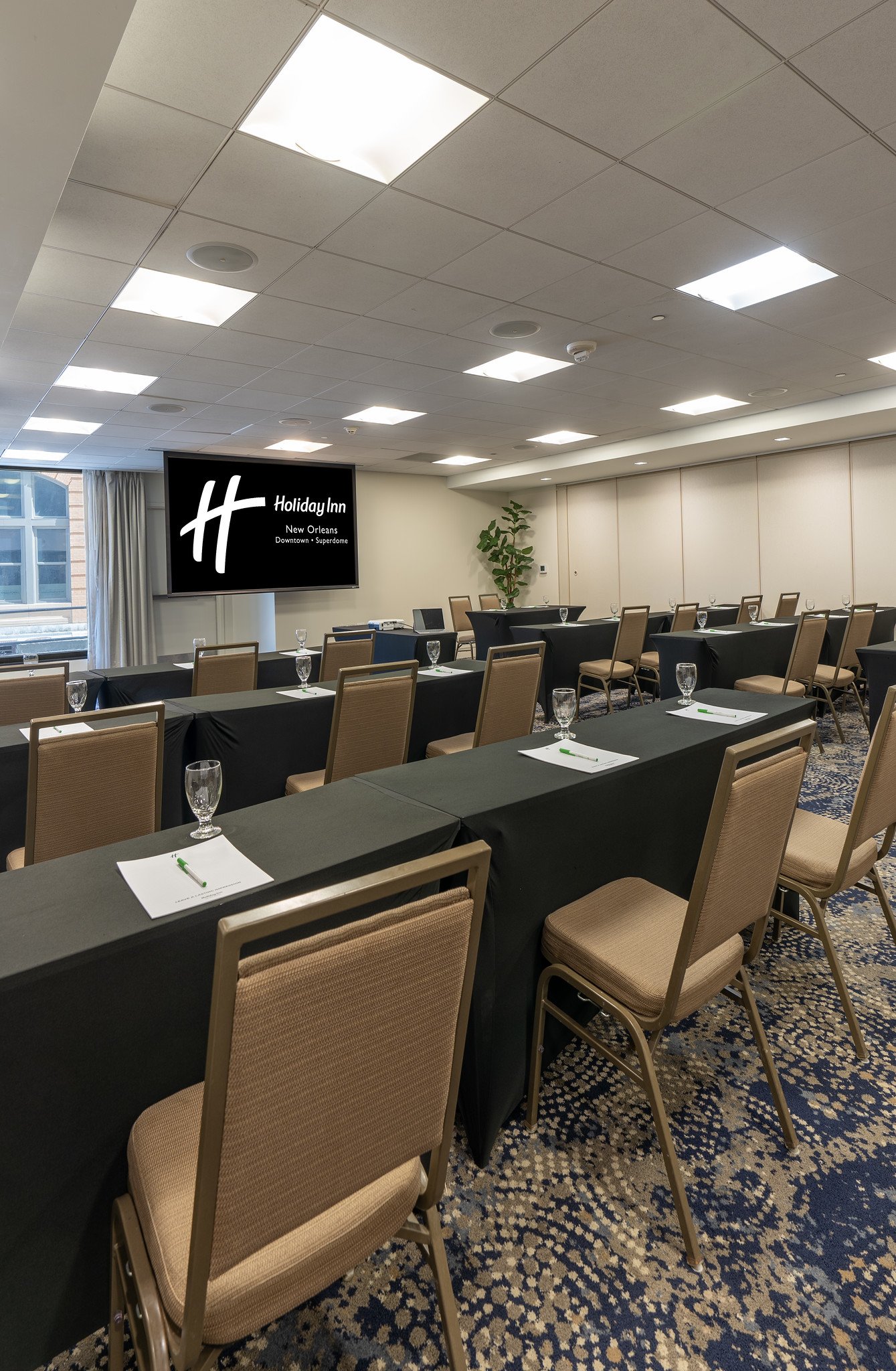 A conference room at Holiday Inn New Orleans Downtown Superdome with rows of tables and chairs, notebooks and pens placed on tables, blank projection screen, and indoor plant for decor.