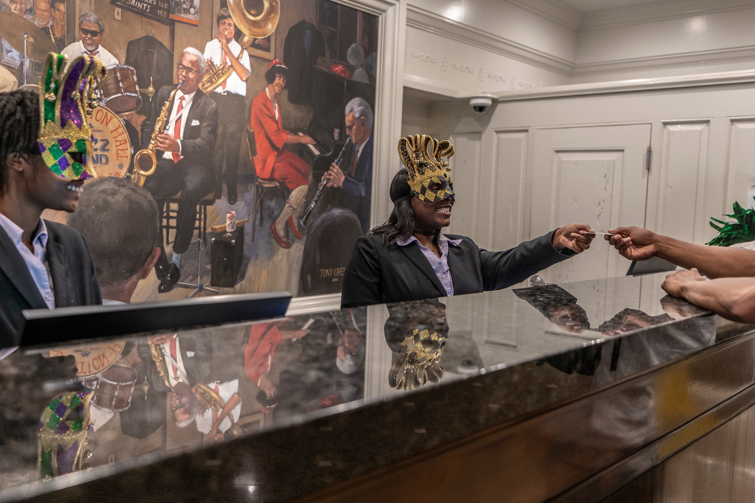Hotel reception with staff wearing Mardi Gras masks handing a card to a guest, mural of jazz musicians in background.