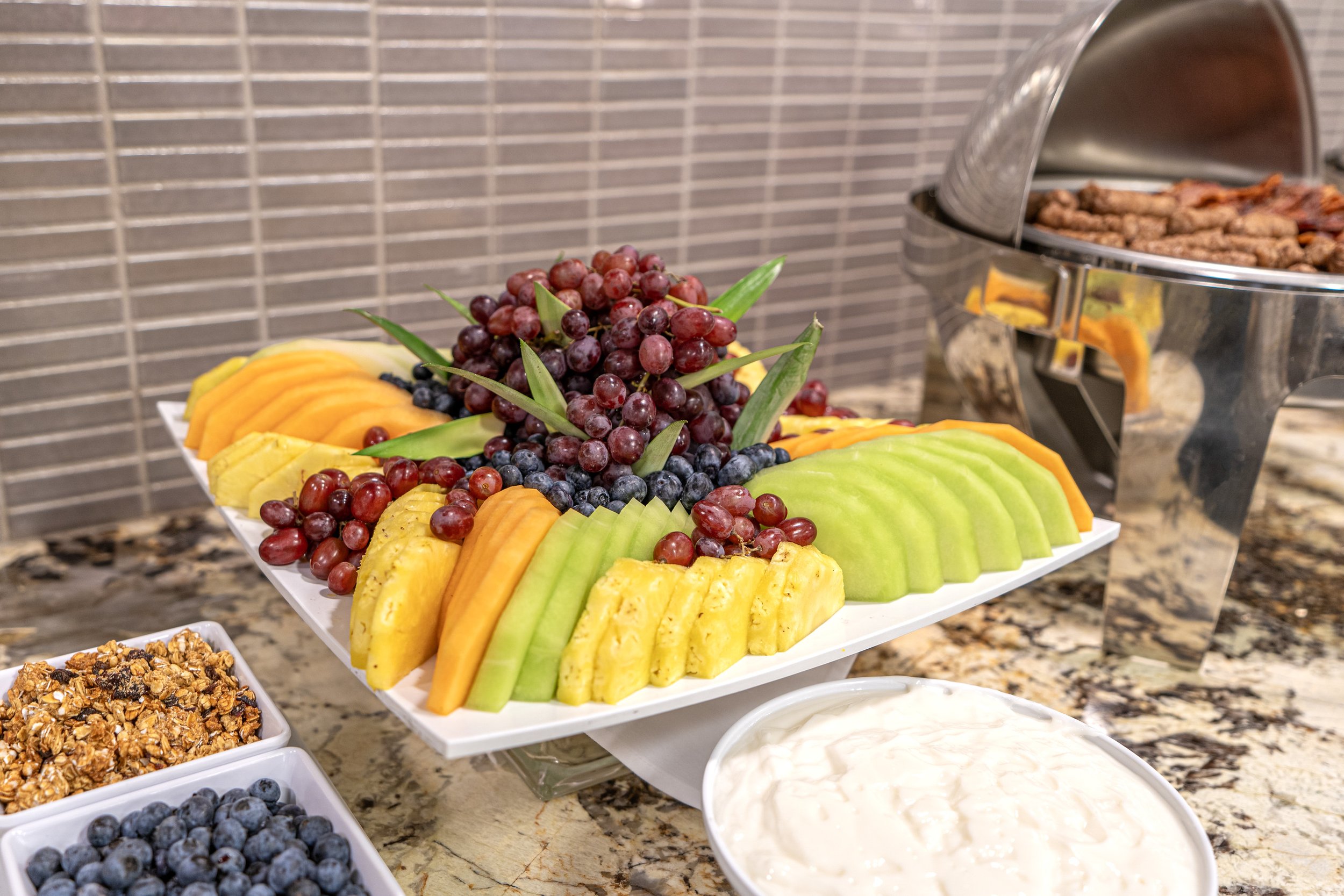 Fruit platter with sliced melons, pineapples, grapes, and blueberries, alongside granola and a bowl of yogurt.