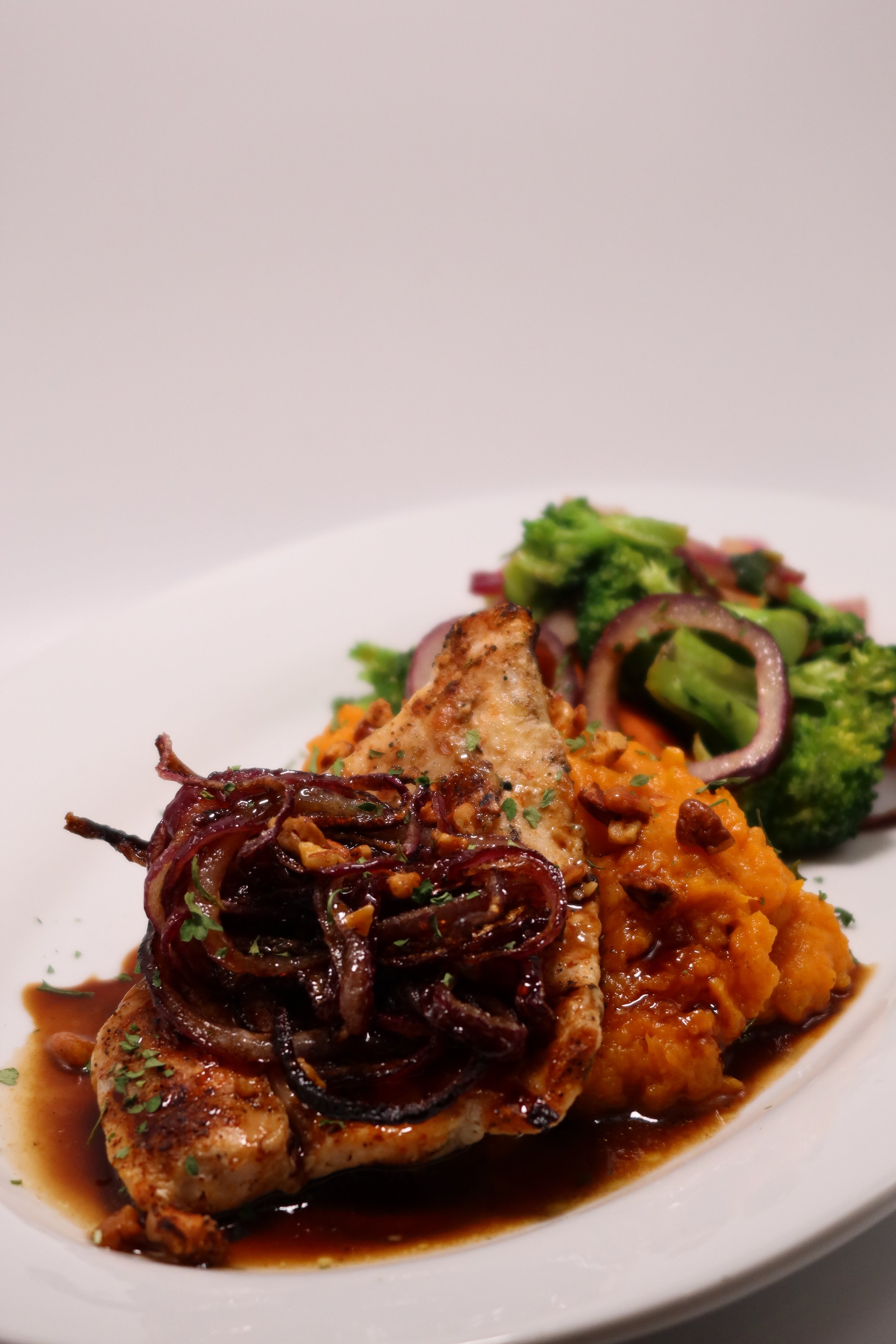 Grilled chicken breast with caramelized onions, mashed sweet potatoes, and steamed vegetables on a white plate.