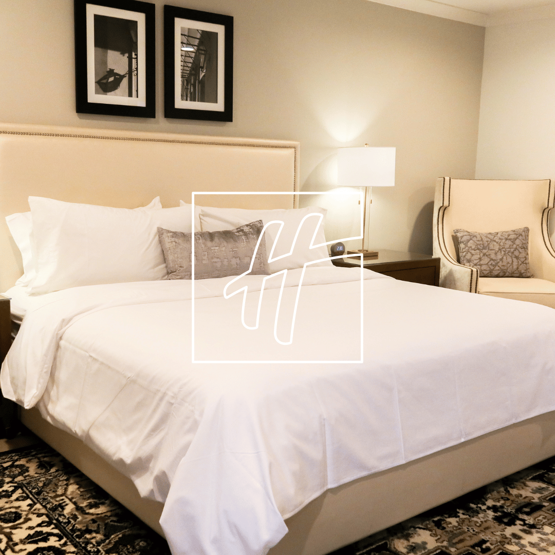 Modern hotel room with a neatly made bed, white linens, decorative pillows, bedside table with lamp, an upholstered chair, and framed artwork on the wall.