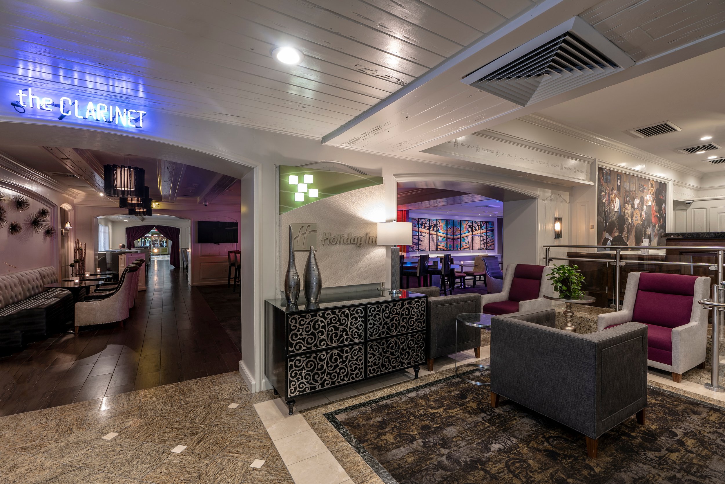 Interior of a hotel lobby with modern decor at Holiday Inn, featuring seating area with purple and gray chairs, decorative cabinet, and neon sign for 'the CLARINET' bar.